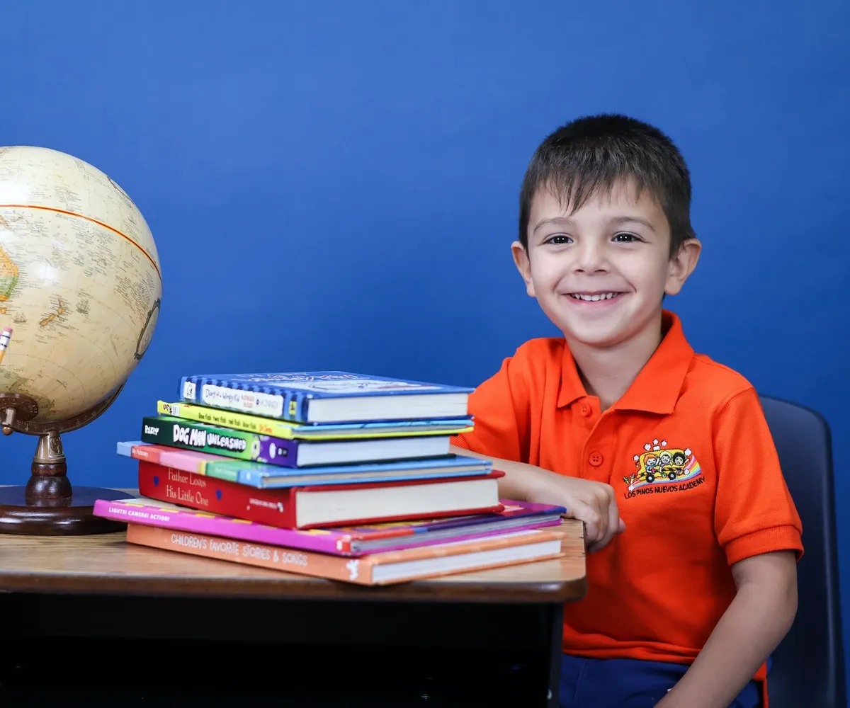 Niño de VPK sonriendo en su pupitre junto a un globo terráqueo y una pila de libros de colores